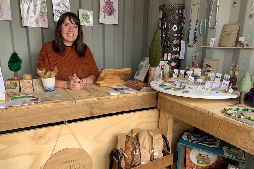 A woman smiles behind a wooden bench displaying jewellery and other items.