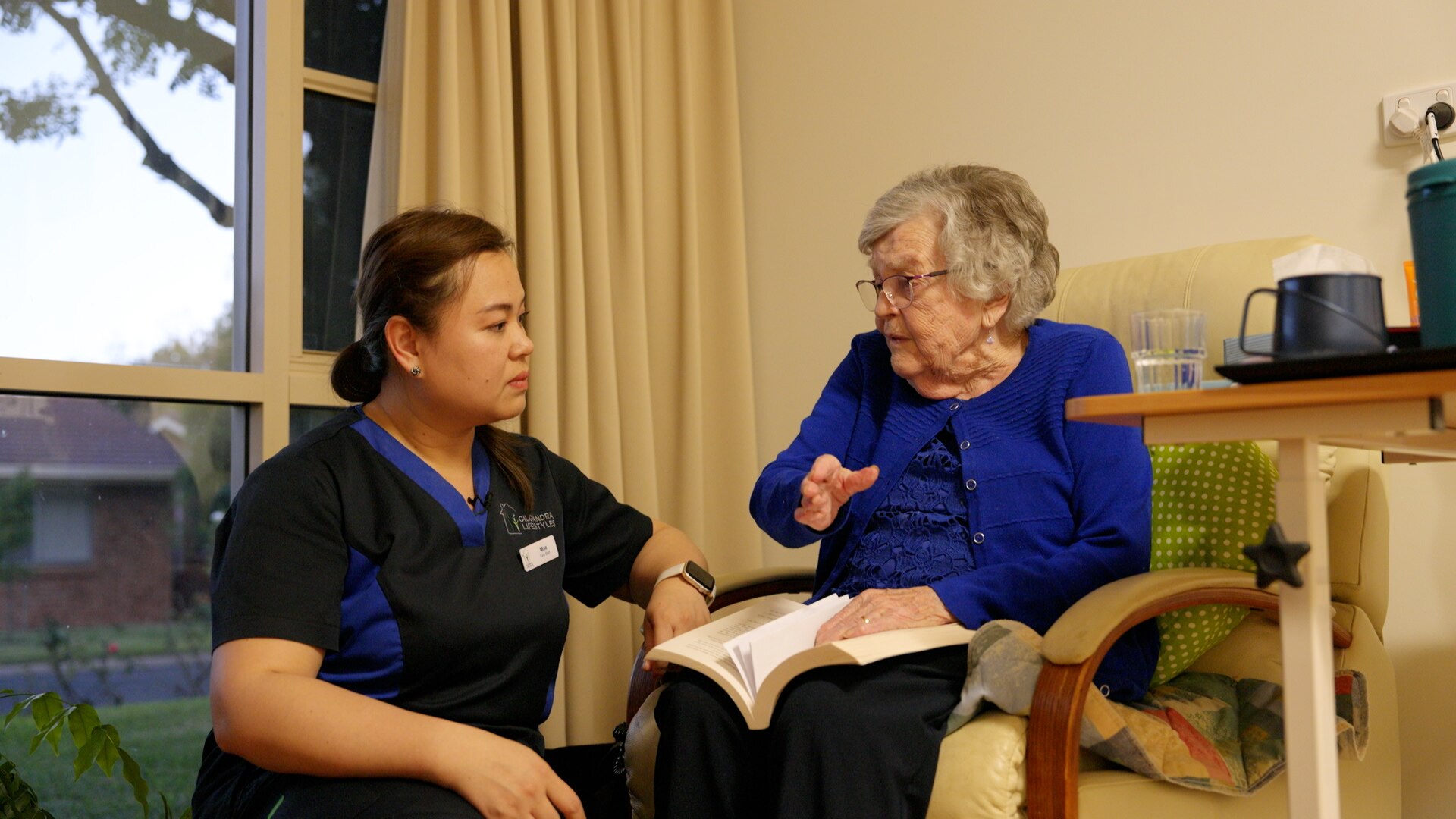 Female healthcare worker squats next to seated elderly woman in aged care facility who is holding a book.