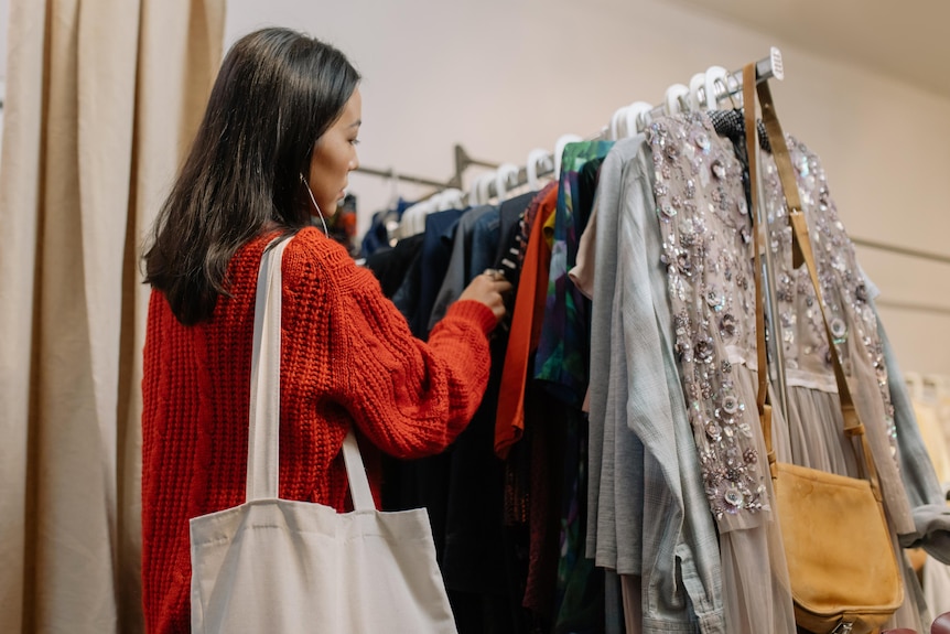 A woman wearing a red knitted jumper and white tote bag looks through rack of clothing in store