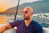 Man wearing sunglasses sits in boat staring out at water with serious expression. 