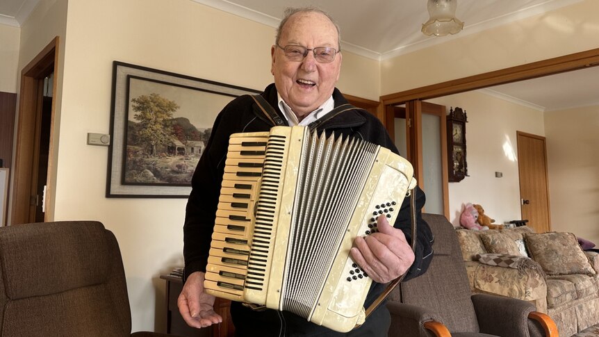 A man chuckles while holding up an ivory white piano accordion in a living room 