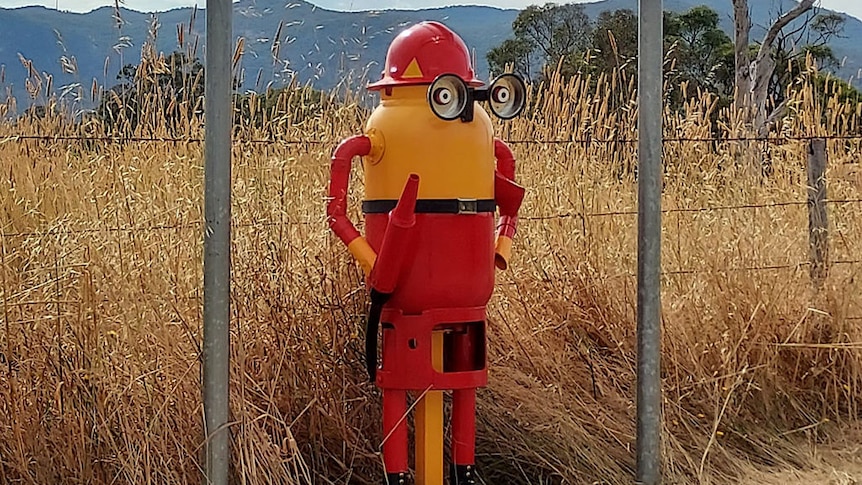A metal sculpture of a yellow minion dressed as a fire fighter stands beside the road with a field behind him