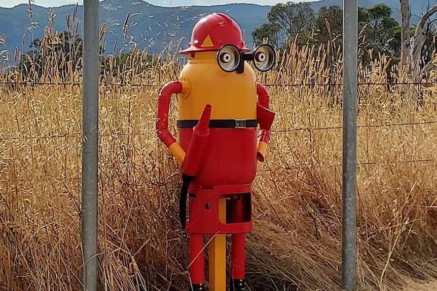 A metal sculpture of a yellow minion dressed as a fire fighter stands beside the road with a field behind him