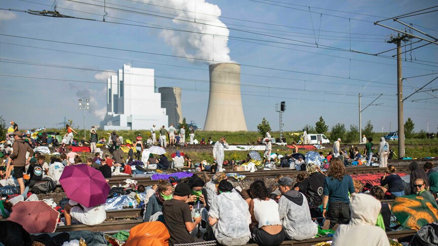 Activists block the tracks of the coal transport railway as smoke rises from an industrial chimney in the background.