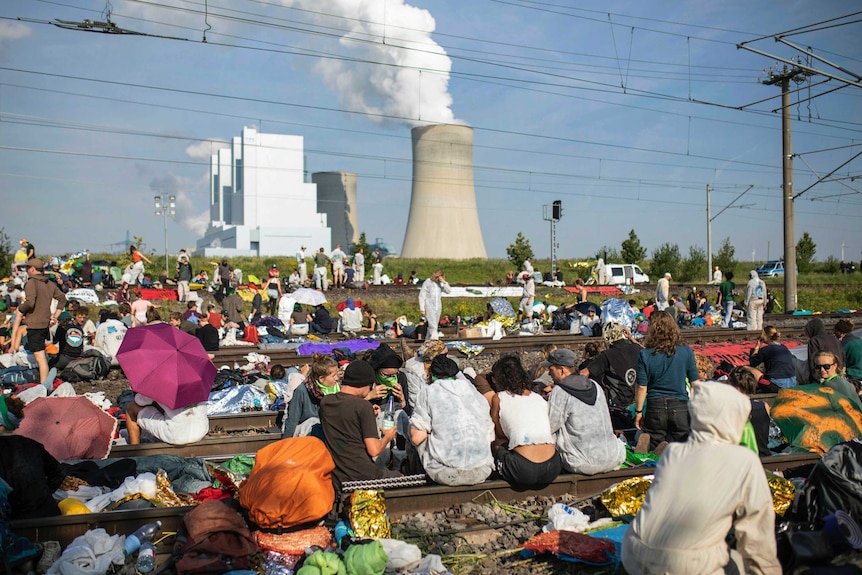 Activists block the tracks of the coal transport railway as smoke rises from an industrial chimney in the background.