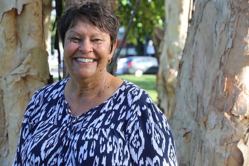 Lynelle stands in front of Eucalyptus trees wearing a white and blue patterned top.