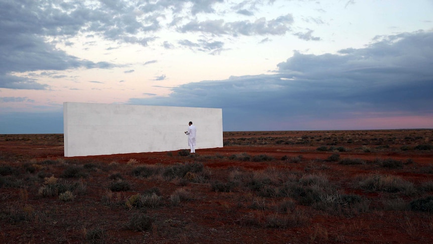 A man dressed in white stands in front of a white wall in a desert setting at sun set.