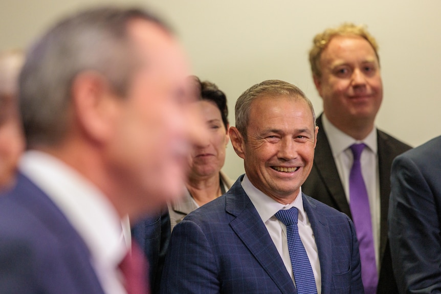 A man in a suit and tie smiles broadly while watching another man in a suit and tie speak. 