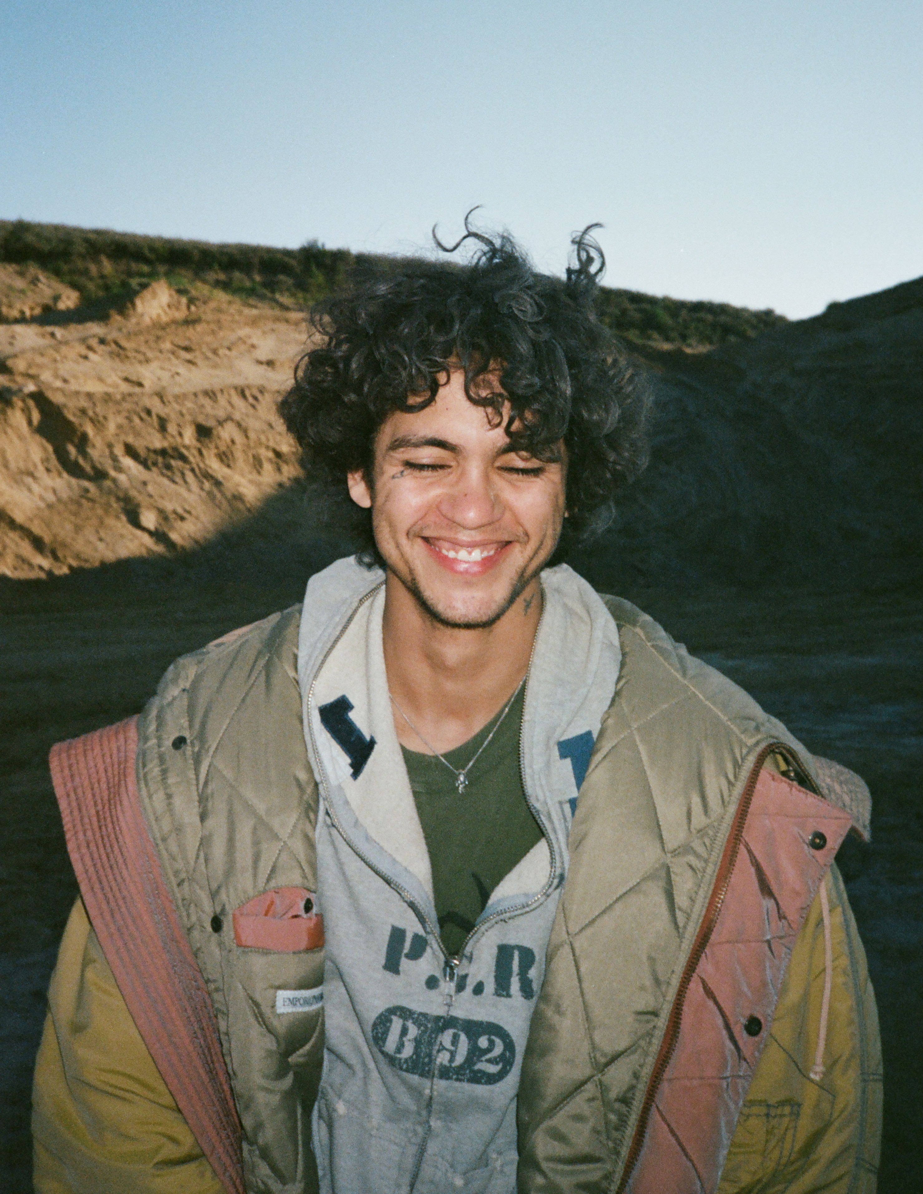 A photo of Dominic Fike smiling with his eyes closed, wearing a hoodie.