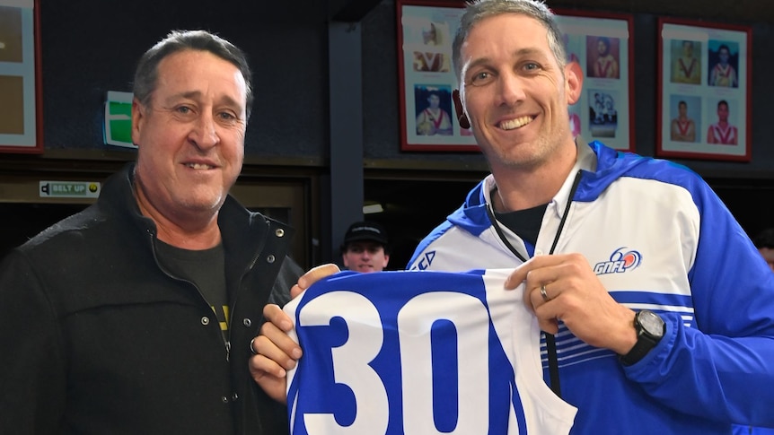 Two men smiling as one holds a football jersey up with number 30 