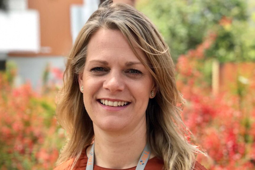 A blonde middle aged woman with fair hair in an orange top and ID badge smiles at camera in park.