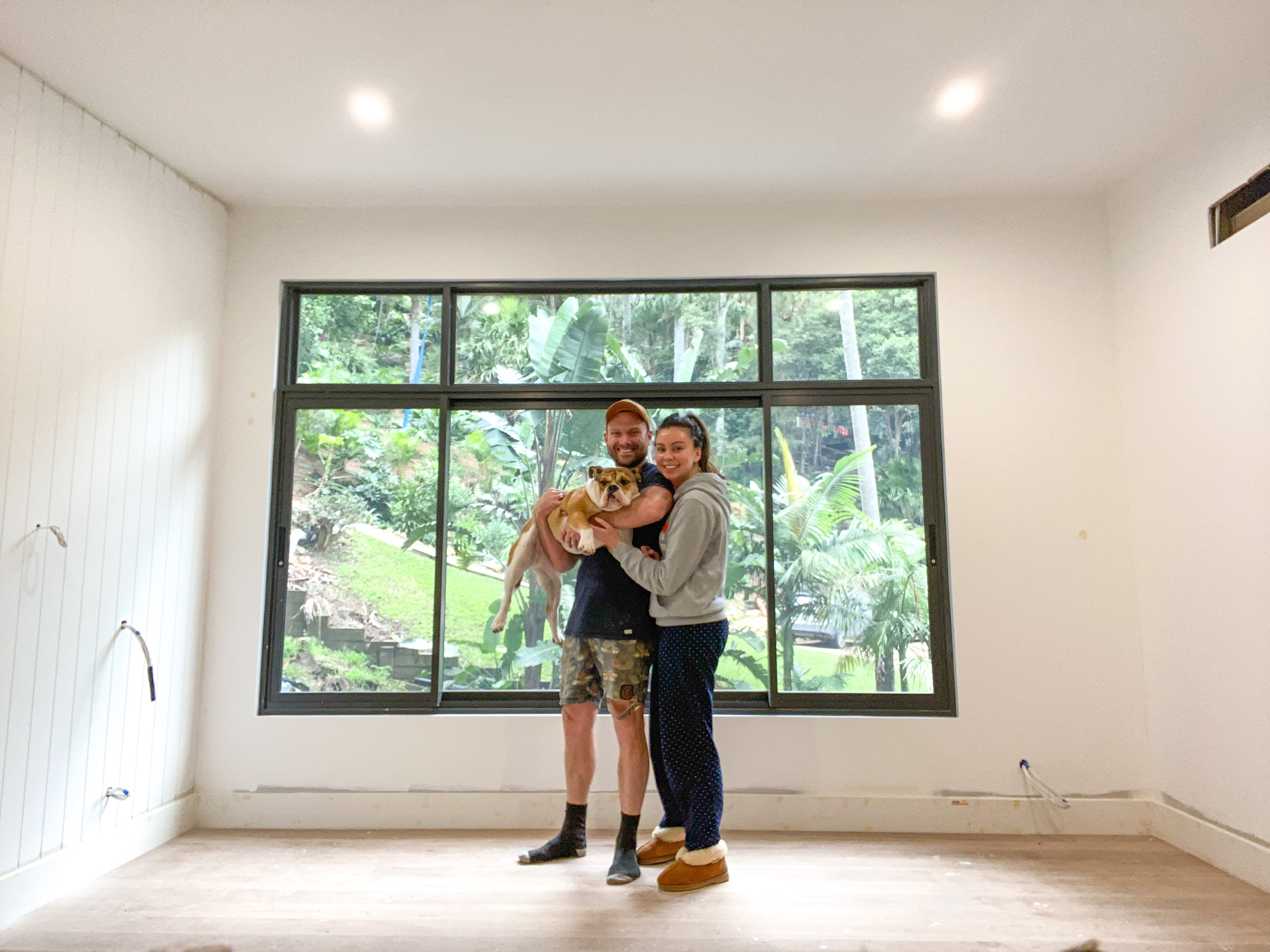 Tamiko Gleeson and her husband Daniel stand in an empty room under construction. They hold their dog and smile at the camera