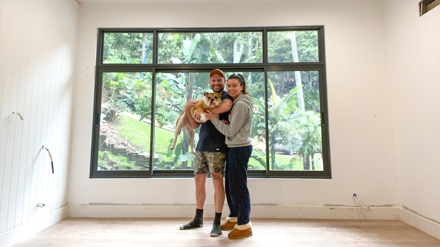 Tamiko Gleeson and her husband Daniel stand in an empty room under construction. They hold their dog and smile at the camera