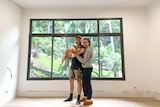 Tamiko Gleeson and her husband Daniel stand in an empty room under construction. They hold their dog and smile at the camera