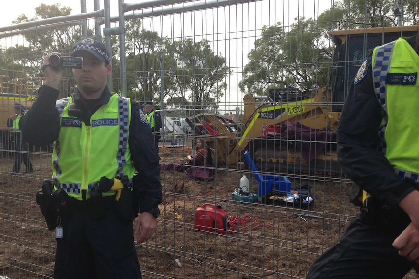 A woman sits in front of a bulldozer behind a fence, with police officers nearby.
