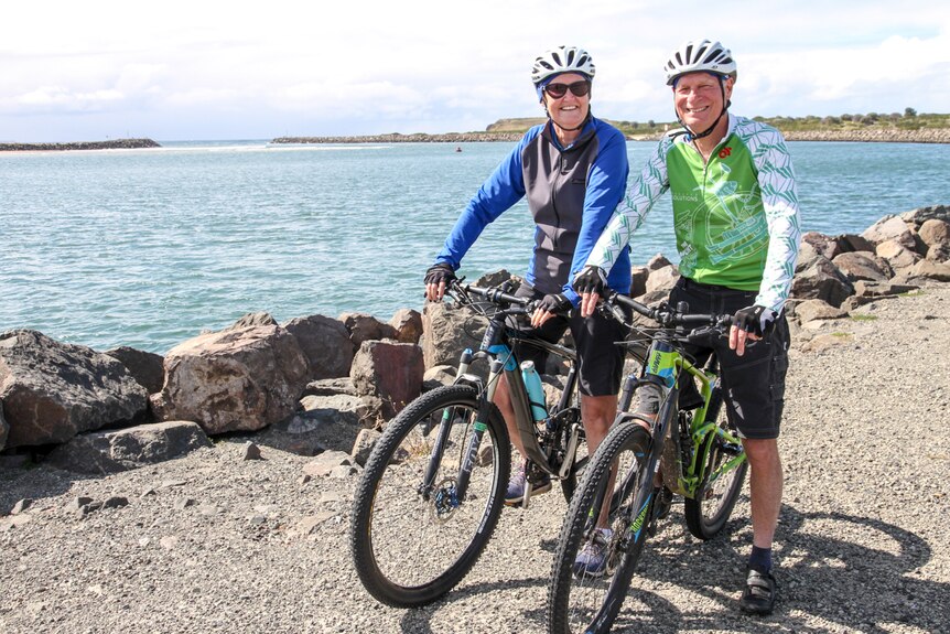 An older couple in cycling gear sit on their bikes in front of a waterway with big smiles on their faces.