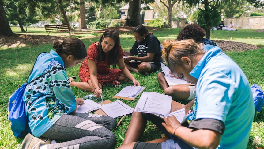 A group of Aboriginal people sitting on grass reading.