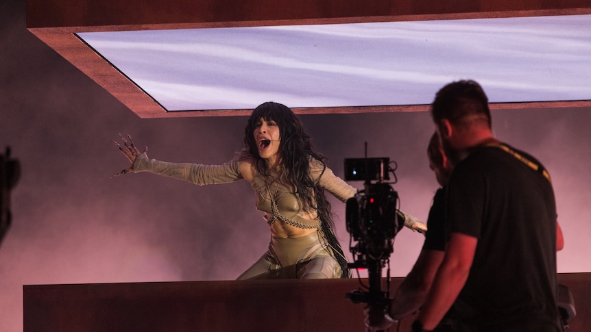 Loreen performing on stage with hands out and a camera in front of her.