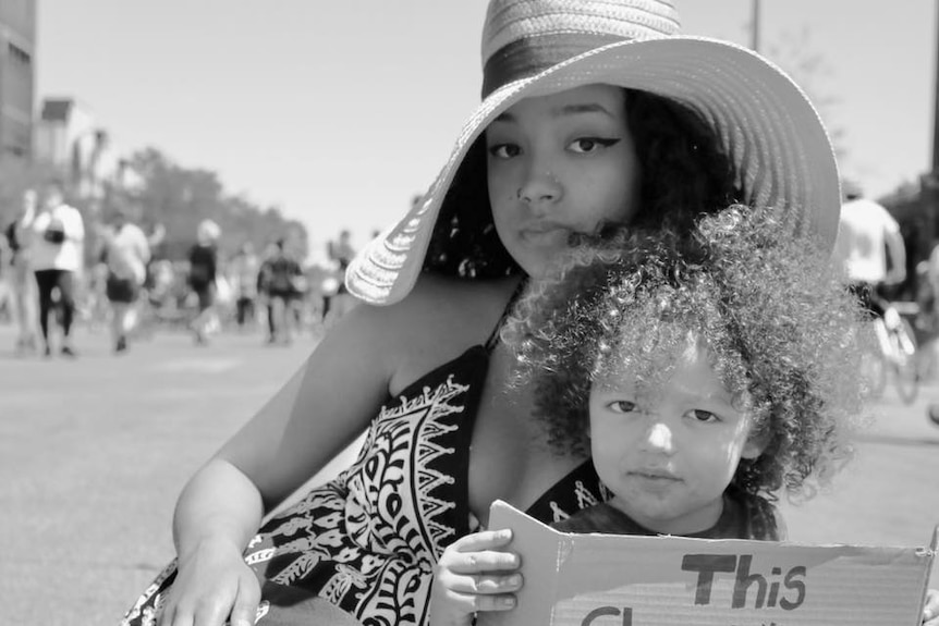 A woman in a sunhat next to a toddler who is holding a sign called 'This shouldn't be my reality!"
