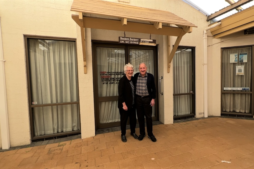 An older man and a woman stand on pavers outside an office, which has a small wooden sign above