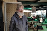 Older man with grey lond beard looks into the distance through glasses in a carport