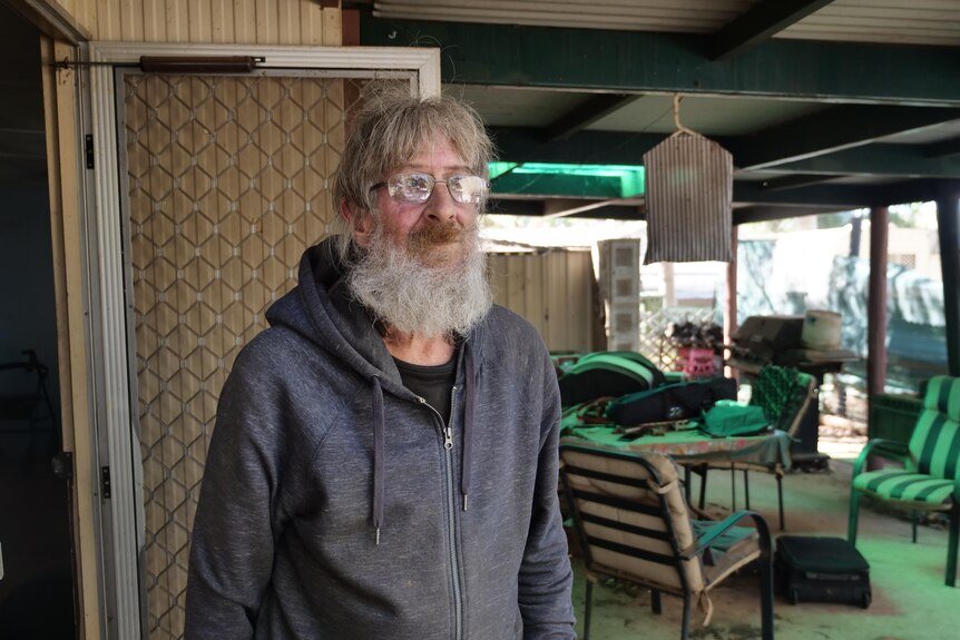 Older man with grey lond beard looks into the distance through glasses in a carport