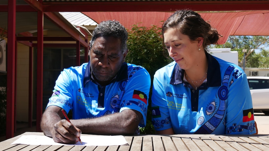 A man and a woman sit at a wooden table outdoors, both wearing blue shirts. The man is drawing on a white piece of paper. 