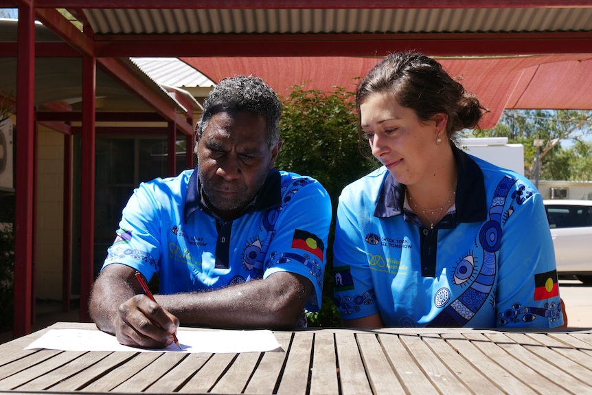 A man and a woman sit at a wooden table outdoors, both wearing blue shirts. The man is drawing on a white piece of paper. 