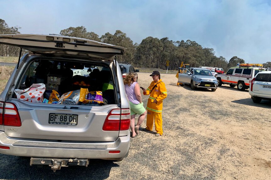 Reporter in yellow fire gear interviewing woman leaning on car parked by road near emergency vehicles.