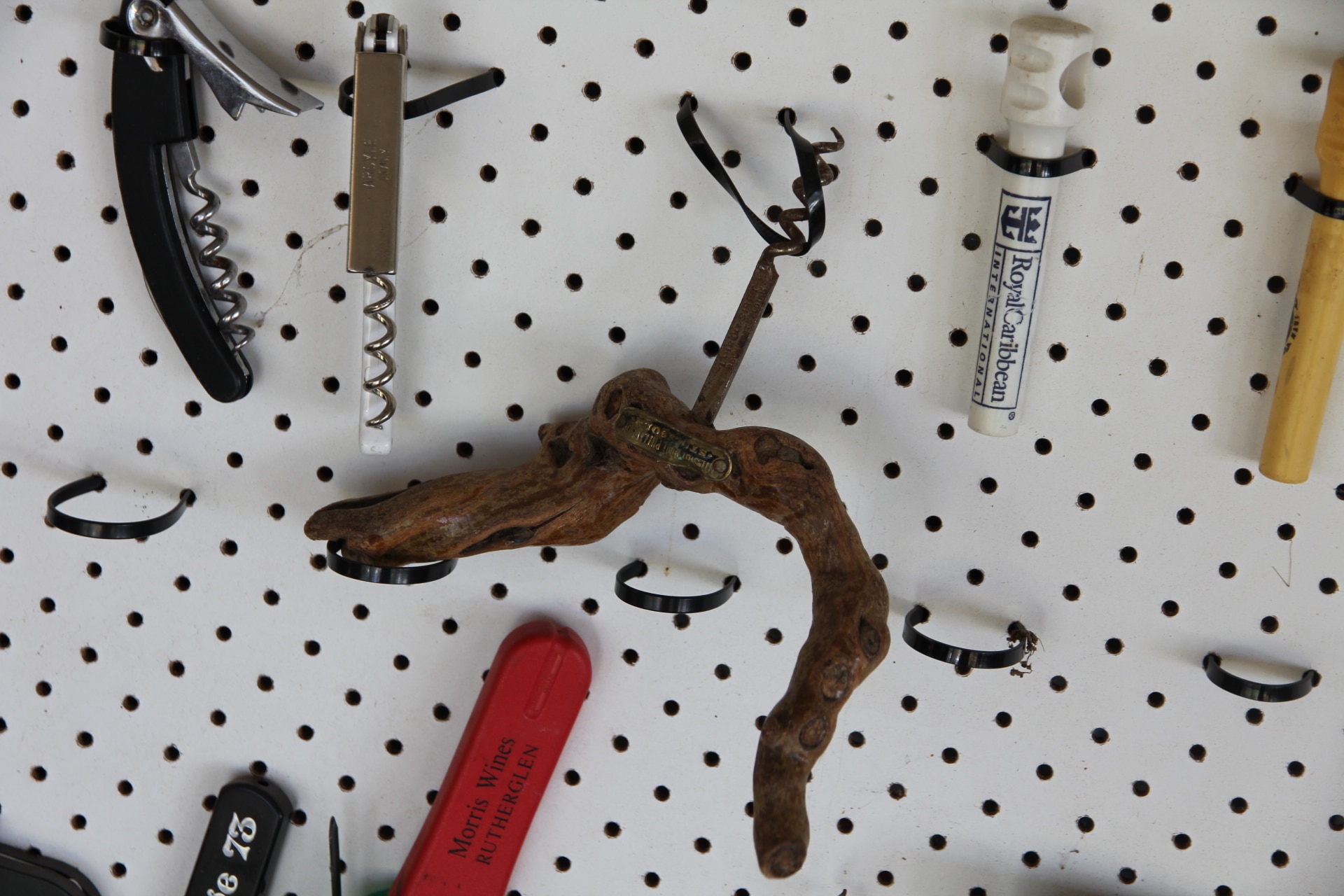 Different corkscrews hanging on a white board. One in the centre has a twisted piece of wood as a handle.