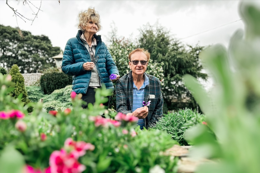 An older woman wearing a blue puffer jacket stands next to a man wearing a plaid coat and sunglasses in a garden.