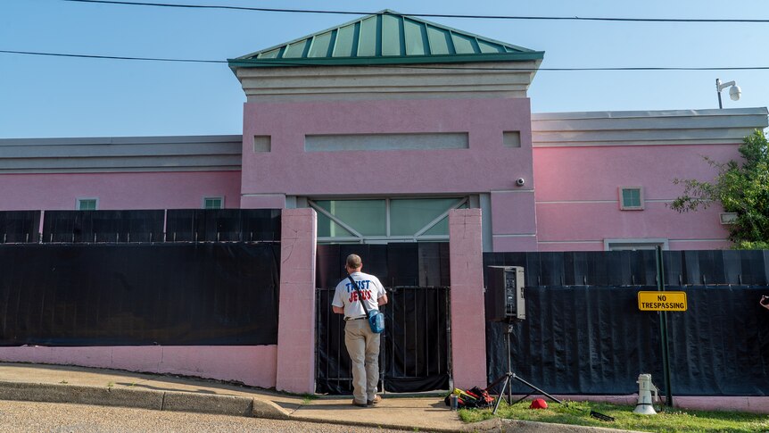 A man wearing a shirt with TRUST JESUS written on it stands in front of a black fence guarding a pink brick building