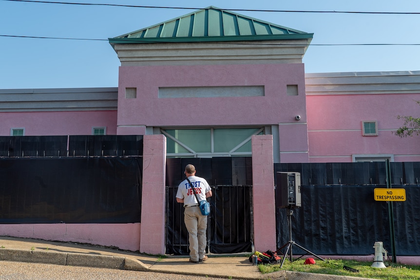 A man wearing a shirt with TRUST JESUS written on it stands in front of a black fence guarding a pink brick building