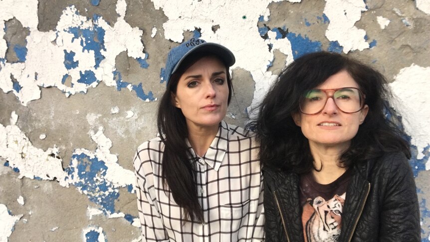 Colour photograph of sisters Dominique and Dan Angeloro of art collective Soda_Jerk standing in front of a distressed wall.