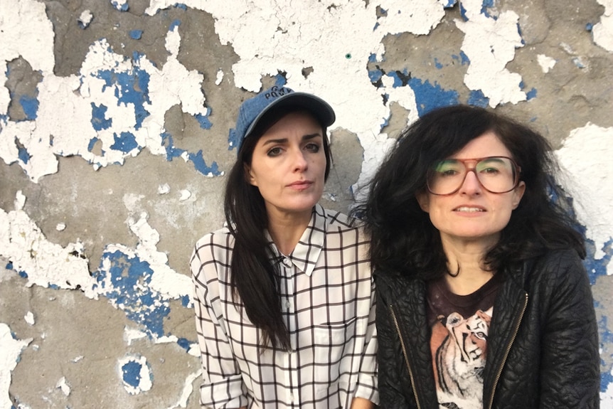 Colour photograph of sisters Dominique and Dan Angeloro of art collective Soda_Jerk standing in front of a distressed wall.