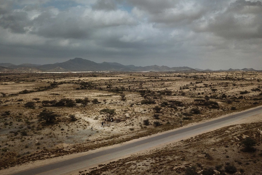 A group of people walk across a desert-like landscape with mountains in the distance.