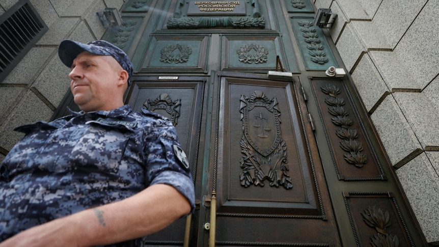 A man in a blue military uniform stands in front of a large wooden door to a court. 