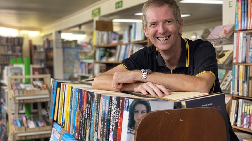 A man leaning on a book case, smiling, with book shelves in the background