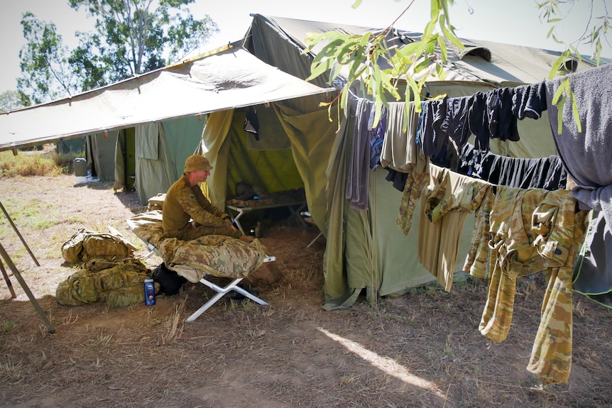 A soldier sits on a stretcher outside a tent with laundry hanging on a makeshift washing line 