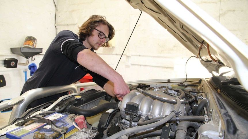 A teenage boy works on a car in a mechanical workshop and looks under the hood.