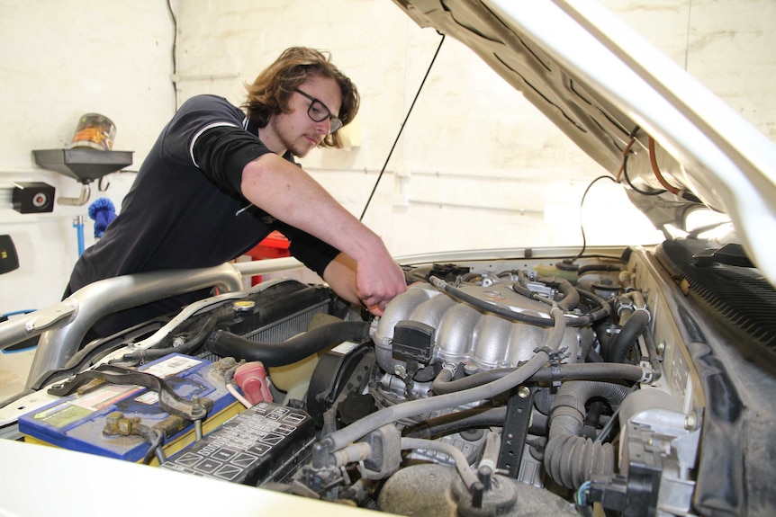 A teenage boy works on a car in a mechanical workshop and looks under the hood.