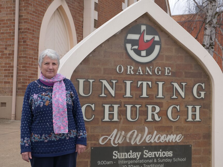 Woman with a pink scarf and blue sweater standing in front of church next to an Orange Uniting church sign