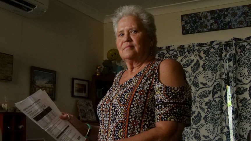 An elderly woman stands in her living room, holding some paperwork and looking concerned.