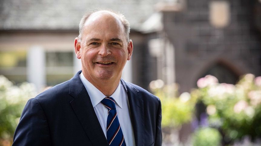 Philip Grutzner smiles at the camera wearing a suit and standing in front a stone building and garden.