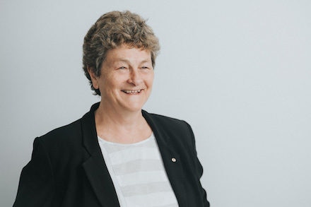 Against a grey background, a short-haired woman from the torso up stands, wearing black jacket and white shirt, smiling widely.