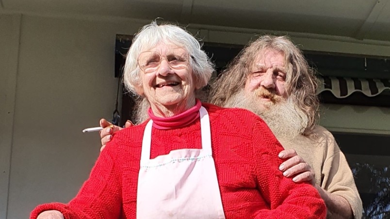 Jock standing behind Iris with arms around her outside house.