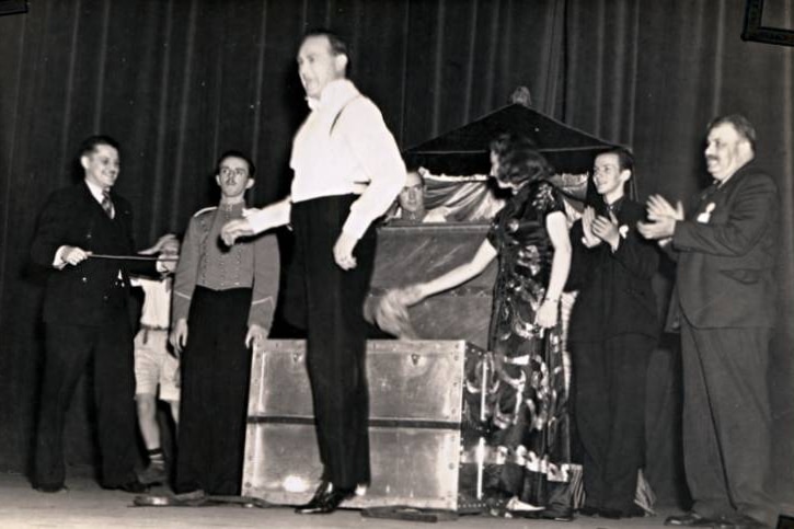 A man jumps up in front of a steel trunk on stage with five people standing around and applauding