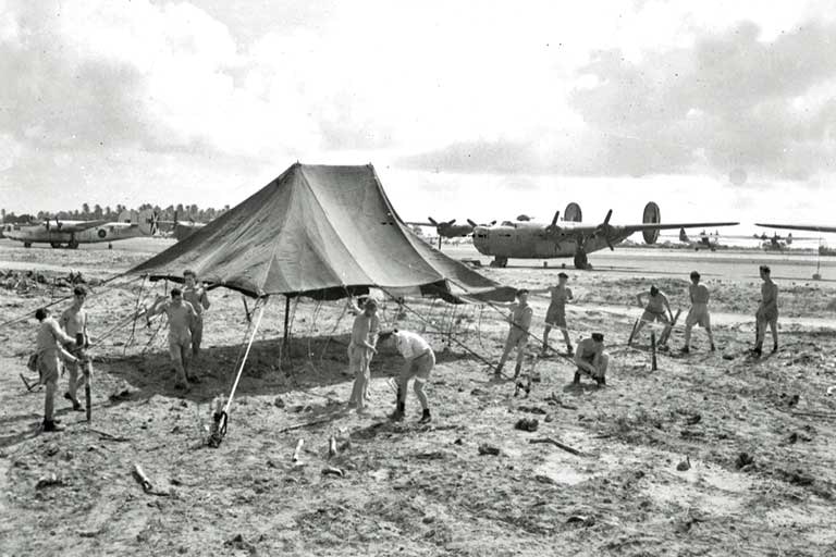 A black and white photo of men building a large tent next to a row of parked aircraft.