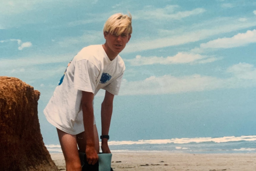 An old photo of a blonde-haired boy putting a wetsuit on at the beach.