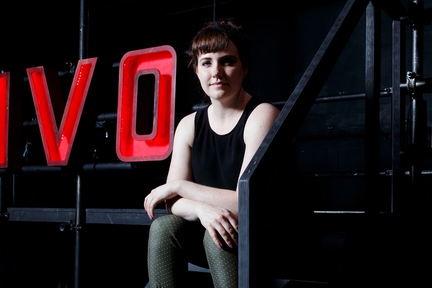 Young woman in black tank top and army-green pants sitting on set with large light-up letters spelling TIVOLI behind her in red.